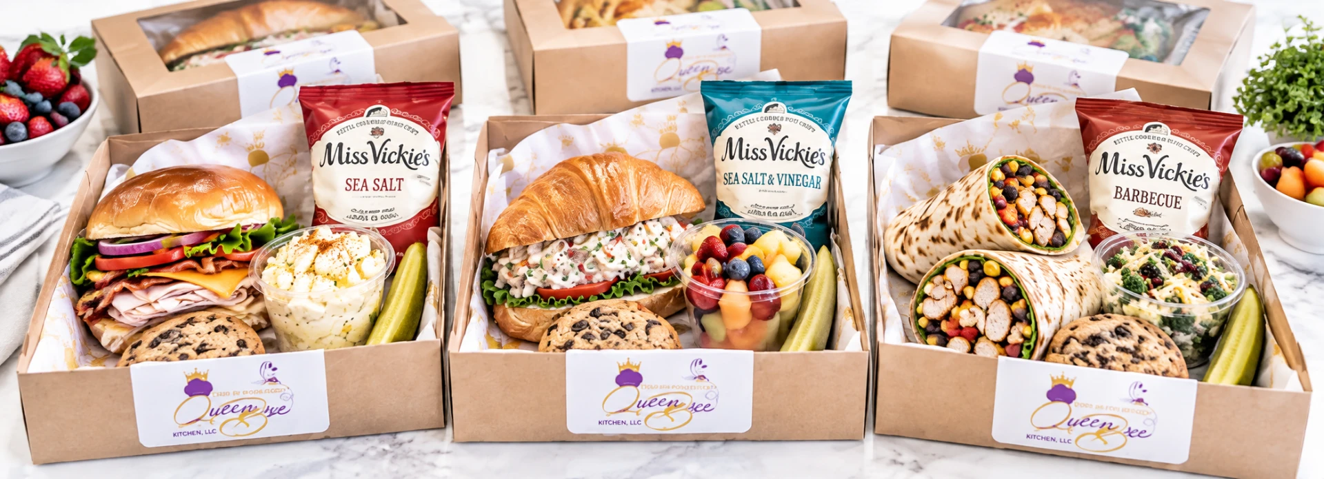 Three Queen Bee Kitchen boxed lunches — turkey club, chicken salad croissant, and southwest chicken wrap — with chips, fruit, and pickles on a conference table